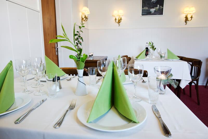 A cozy restaurant dining room features elegantly set tables with white linens, green napkins, and simple floral centerpieces, creating an inviting atmosphere perfect for intimate gatherings or romantic dinners.