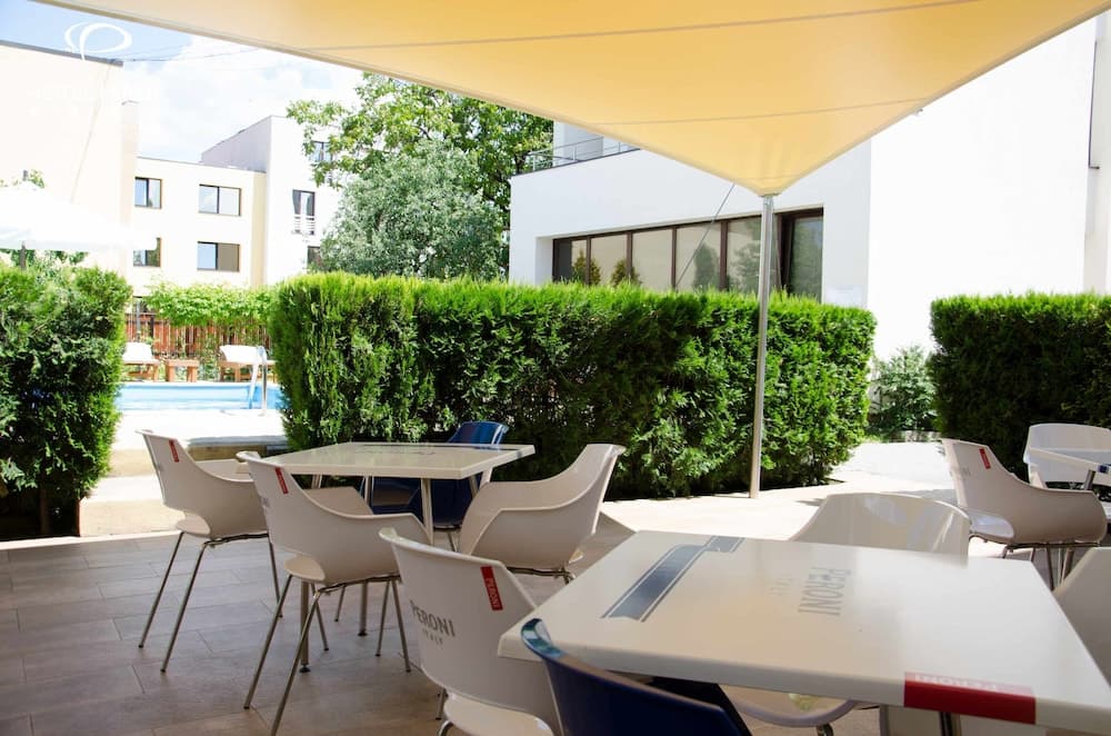 A serene outdoor café patio features white tables and chairs under a large canopy, surrounded by lush green hedges and a glimpse of a swimming pool, ideal for travelers seeking a relaxing dining spot near modern buildings.