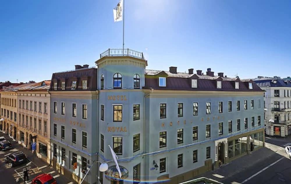 A charming blue Hotel Royal stands proudly in a bustling urban square under a clear blue sky, featuring classic architecture and a waving flag, ideal for travelers seeking a historic stay in a vibrant city center.
