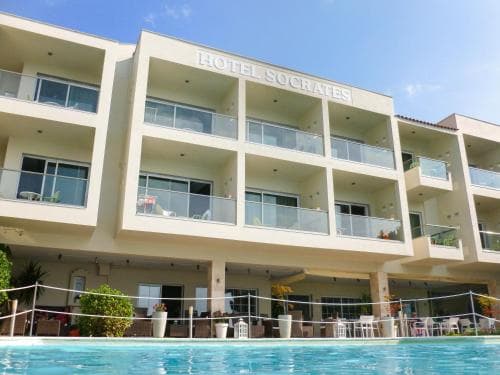 A sunny day at a modern hotel featuring "Hotel Socrates" signage, with a clear blue sky, balconies overlooking a vibrant pool area, and lush greenery, ideal for travelers seeking relaxation and leisure.