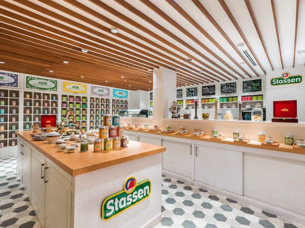 A cozy and inviting Stassen store features neatly arranged shelves of colorful jars, a central display counter with tea sets, and a charming hexagonal-tiled floor, perfect for a delightful shopping experience.