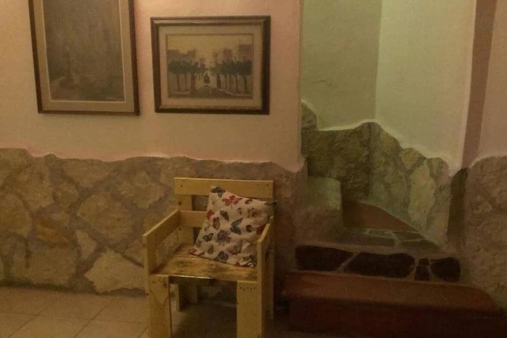 A cozy indoor nook features rustic stone walls, framed artwork, a wooden chair with a floral pillow, and a cushioned bench, ideal for travelers seeking a quaint and serene resting spot.