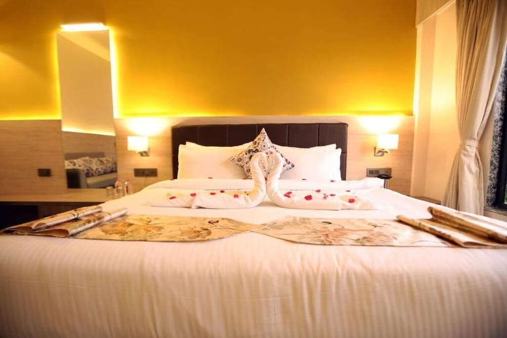A cozy hotel room with a warmly lit yellow accent wall features a neatly made bed adorned with swan-shaped towels and rose petals, ideal for romantic getaways or luxurious stays.