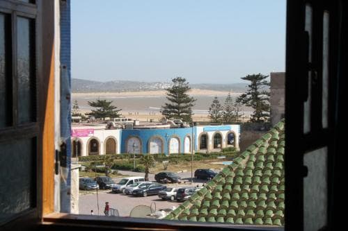 A serene coastal view framed by an open window showcases a vibrant blue building with arched windows, parked cars, and lush greenery, ideal for travelers seeking a tranquil seaside retreat.