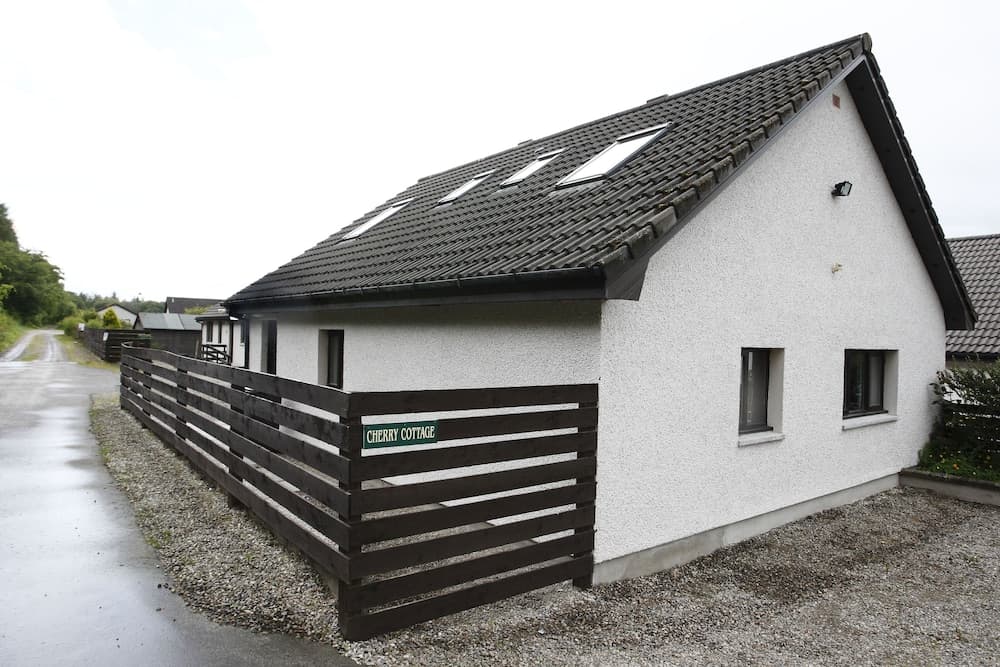 A cozy white cottage named "Cherry Cottage" with a dark wooden fence stands invitingly on a quiet gravel path, ideal for travelers seeking a peaceful rural retreat.
