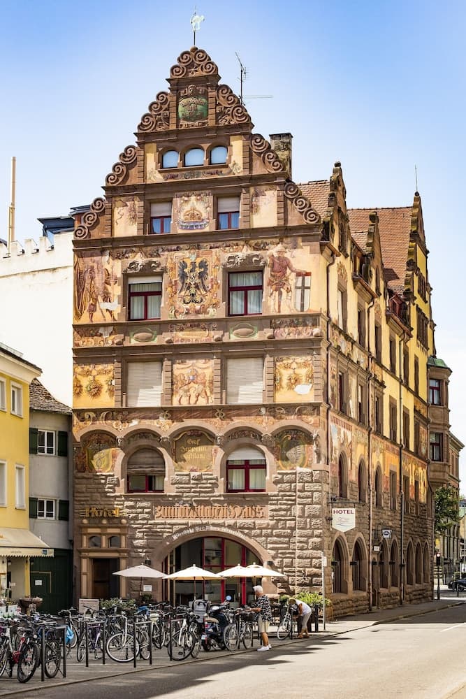 A charming European town square features a historic hotel adorned with intricate frescoes and ornate architecture, bustling with bicycles and outdoor seating, ideal for travelers seeking cultural immersion and picturesque photo opportunities.