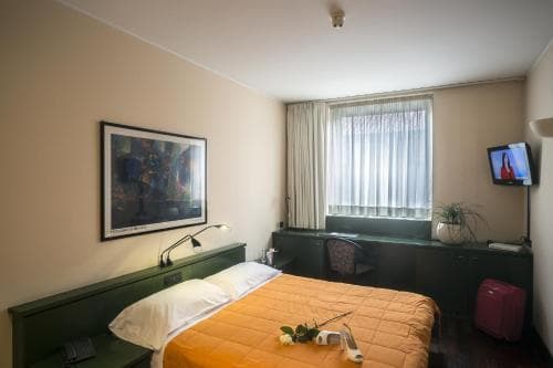 A cozy hotel room features a neatly made bed with an orange blanket, a desk by the window with sheer curtains, a wall-mounted TV, and a vibrant painting, ideal for travelers seeking comfort and simplicity.
