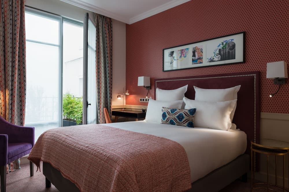A cozy hotel room features a plush bed adorned with white linens and a patterned throw, set against a vibrant red wall with modern artwork, complemented by a purple armchair and a balcony view, ideal for travelers seeking comfort and style.