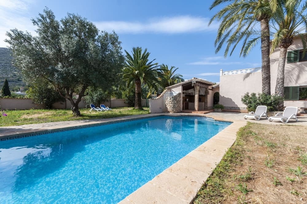 A serene private villa poolside scene features crystal-clear water, lounge chairs, lush palm trees, and olive groves under a bright blue sky, ideal for a relaxing vacation retreat.