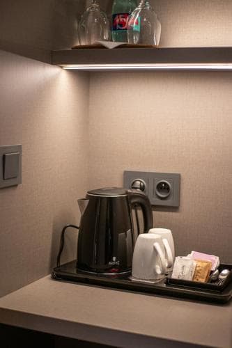 A cozy hotel room corner features a sleek electric kettle, cups, tea packets, and water bottles on a tray, set against soft beige walls with modern lighting, ideal for travelers seeking comfort and convenience.