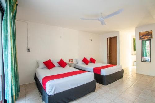 A cozy hotel room with two neatly made beds adorned in red accents, featuring a tiled floor, ceiling fan, and green curtains, ideal for travelers seeking comfort and simplicity.