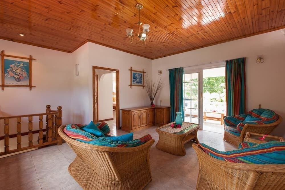 A cozy tropical living room with wicker furniture, vibrant blue and red cushions, wooden ceilings, and large glass doors leading to a balcony, ideal for a relaxing vacation retreat.