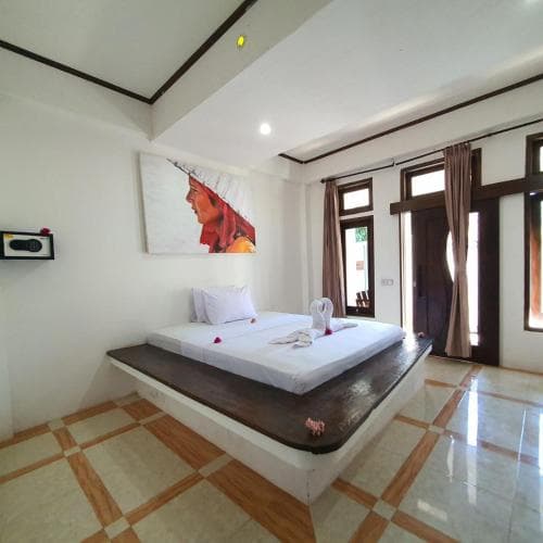 A serene hotel room features a minimalist design with a platform bed adorned with white linens and a towel swan, set against tiled floors and large windows, ideal for travelers seeking a tranquil retreat.