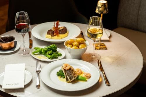 An elegantly set dining table in a sophisticated restaurant features gourmet dishes like seared fish and beef with vibrant vegetables, complemented by fine wine glasses and a luxurious ambiance, perfect for a romantic dinner or a special occasion celebration.