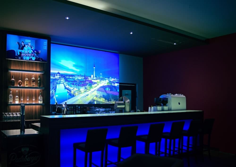 A modern bar with a vibrant blue-lit counter, illuminated shelves of spirits, and a large screen displaying a cityscape creates an inviting atmosphere ideal for travelers seeking a stylish evening spot.