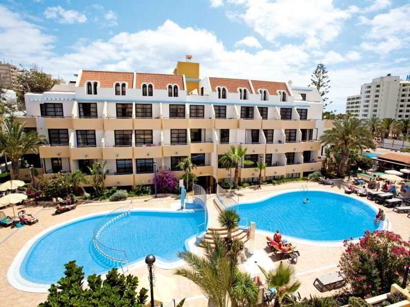 A vibrant resort scene featuring a large swimming pool surrounded by loungers and lush greenery, set against a sunny backdrop with a multi-story hotel building, ideal for family vacations seeking relaxation and leisure.