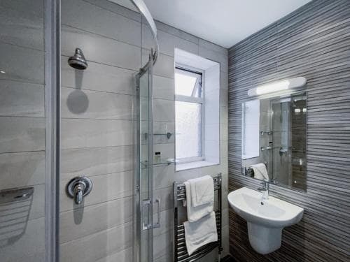A modern bathroom with sleek tiled walls, a glass-enclosed shower, a small window, and a sink with a mirror, ideal for travelers seeking a clean and stylish accommodation space.