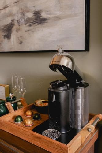 A cozy hotel room corner features a stylish coffee machine on a wooden tray adorned with chocolates, evoking a warm and inviting atmosphere perfect for a relaxing getaway.