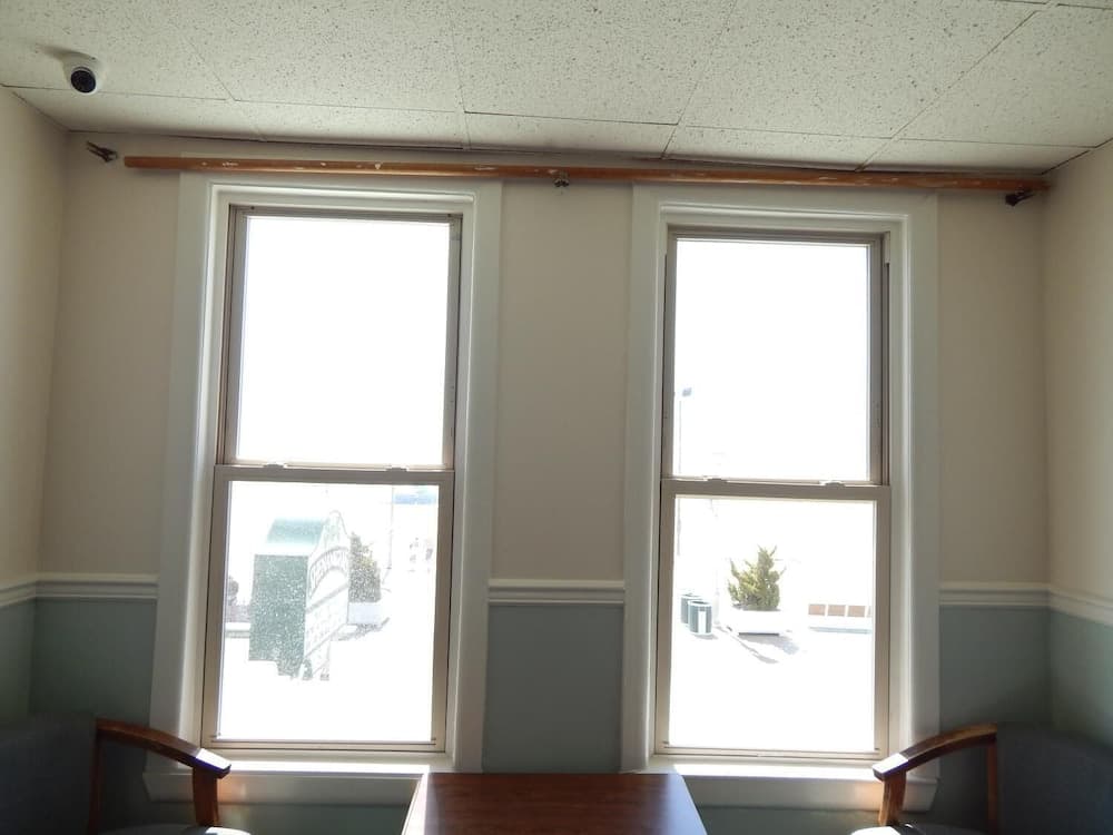 A cozy office space features two large windows with a bright outdoor view, wooden armchairs, and a table, ideal for a serene work-from-home setup or a quiet meeting room ambiance.