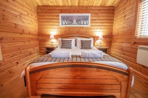 A cozy wooden cabin bedroom with a neatly made bed, warm lighting, and a scenic painting evokes a serene retreat ideal for travelers seeking relaxation in nature.