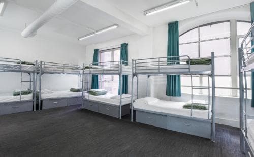 A cozy hostel room with neatly arranged bunk beds, teal curtains, and ample natural light creates a welcoming atmosphere ideal for budget travelers seeking comfortable accommodations.
