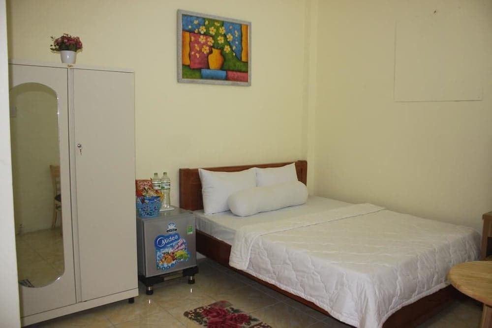 A cozy hotel room features a neatly made bed, a small fridge stocked with snacks, a wardrobe with a mirror, and vibrant wall art, ideal for travelers seeking comfort and convenience.