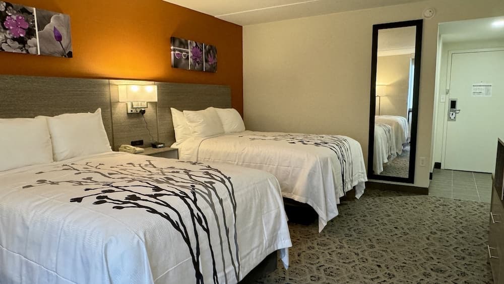 A cozy hotel room features two neatly made beds with floral-patterned bedding, an orange accent wall adorned with purple flower artwork, a full-length mirror, and a warm ambiance ideal for travelers seeking comfort and relaxation.