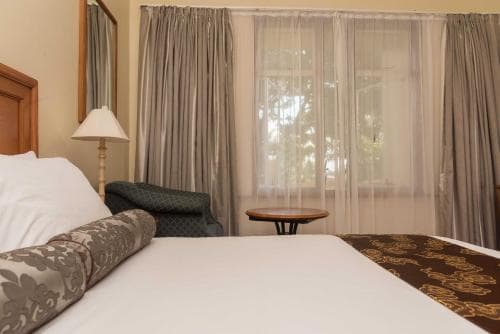 A cozy hotel room features a neatly made bed with patterned linens, soft curtains framing a window view of greenery, and a warm ambiance ideal for travelers seeking relaxation.