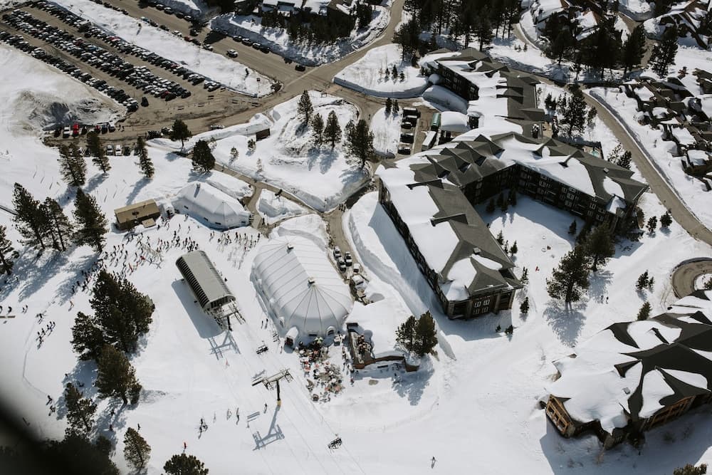 An aerial view captures a bustling winter resort with snow-covered buildings, a large dome structure, and scattered evergreens, ideal for travelers seeking a serene yet lively ski vacation experience.