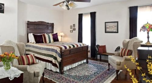 A cozy bedroom with a wooden bed adorned in colorful bedding, surrounded by plush armchairs, a vibrant rug, and floral accents, creates an inviting atmosphere ideal for a relaxing getaway.