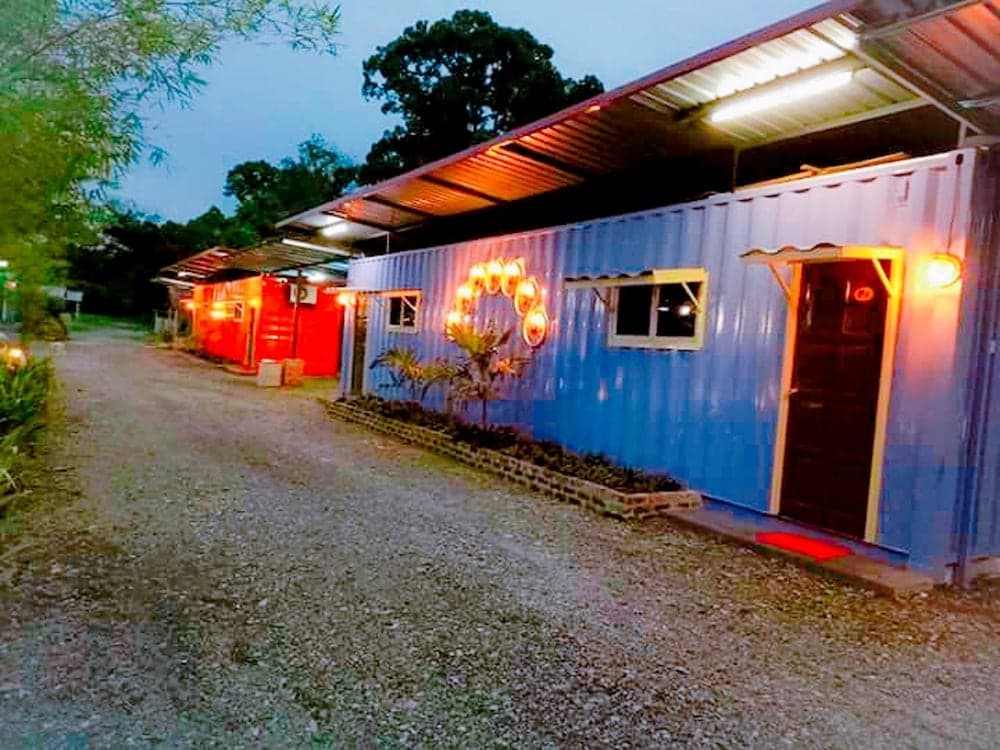 A cozy evening scene at a shipping container village with warmly lit blue and red units, inviting travelers to explore unique accommodations amidst lush greenery.