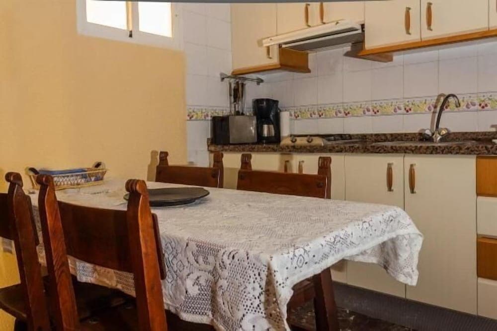 A cozy kitchen with cream cabinets, tiled backsplash, and a wooden dining table draped in a lace tablecloth creates a warm, inviting atmosphere ideal for travelers seeking a homely rental experience.