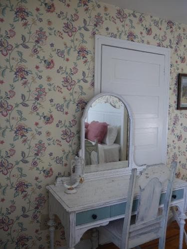 A cozy bedroom features a vintage vanity with a mirror reflecting floral wallpaper, creating a charming and nostalgic atmosphere ideal for a relaxing getaway.