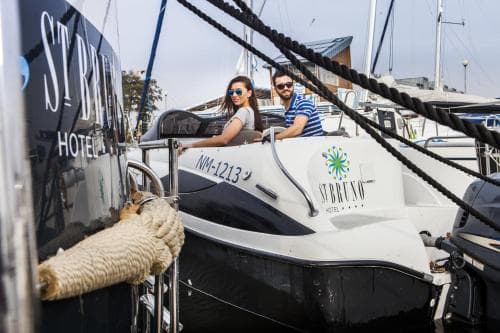 A couple enjoys a sunny day aboard a sleek motorboat docked at a marina, surrounded by other vessels and a hotel sign, ideal for travelers seeking leisure and nautical adventures.