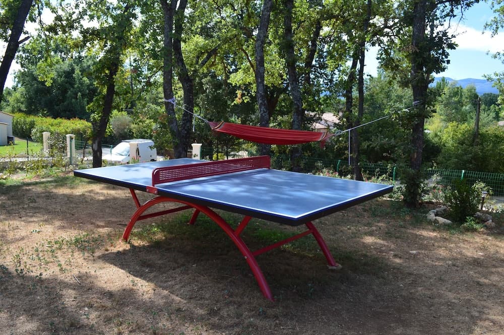 A serene outdoor setting features a red ping pong table under the shade of tall trees, complemented by a hammock and lush greenery, ideal for a relaxing family vacation or recreational retreat.