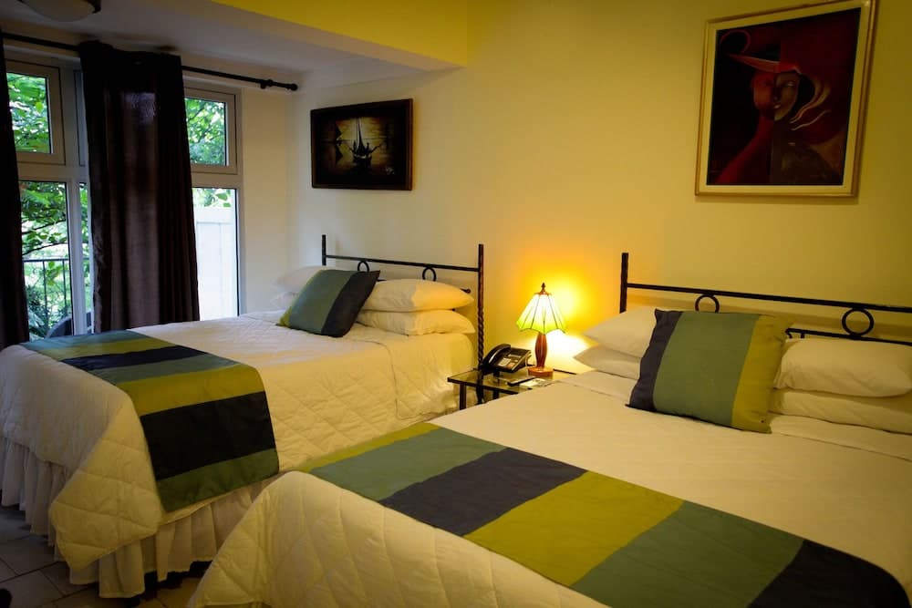 A cozy hotel room with two neatly made beds adorned in green and white bedding, illuminated by a warm lamp beside an artistic wall decor, offers a serene retreat ideal for travelers seeking comfort and relaxation.