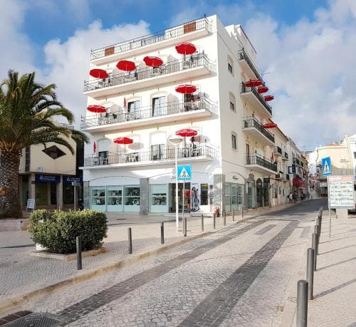 A vibrant street scene in a sunny coastal town features a charming multi-story building with balconies adorned by red umbrellas, inviting travelers to enjoy leisurely strolls and alfresco dining.