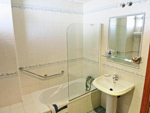 A cozy hotel bathroom features a bathtub with a glass shower screen, a pedestal sink, and a mirrored shelf, creating a clean and functional space ideal for travelers seeking comfort.