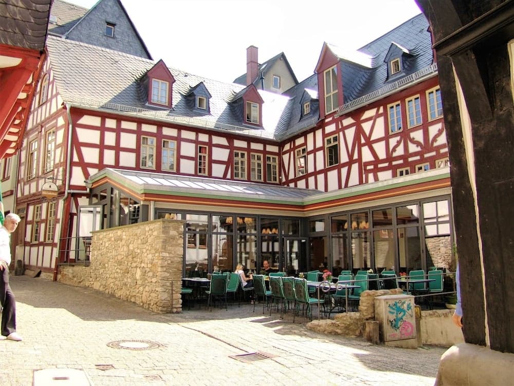 A charming European courtyard features half-timbered buildings, outdoor seating at a café, and cobblestone pathways, ideal for travelers seeking a quaint and relaxing atmosphere.