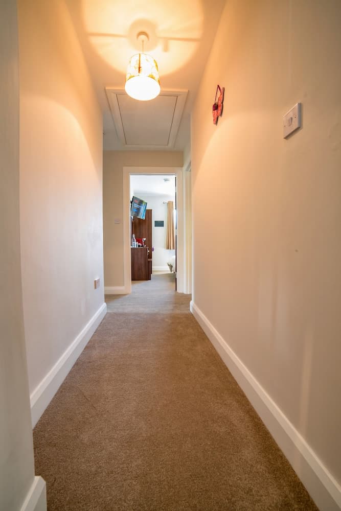 A cozy, warmly lit residential hallway with beige carpeting, white walls, a decorative lamp, and a glimpse into a lived-in room creates an inviting atmosphere ideal for showcasing home interiors in real estate listings.