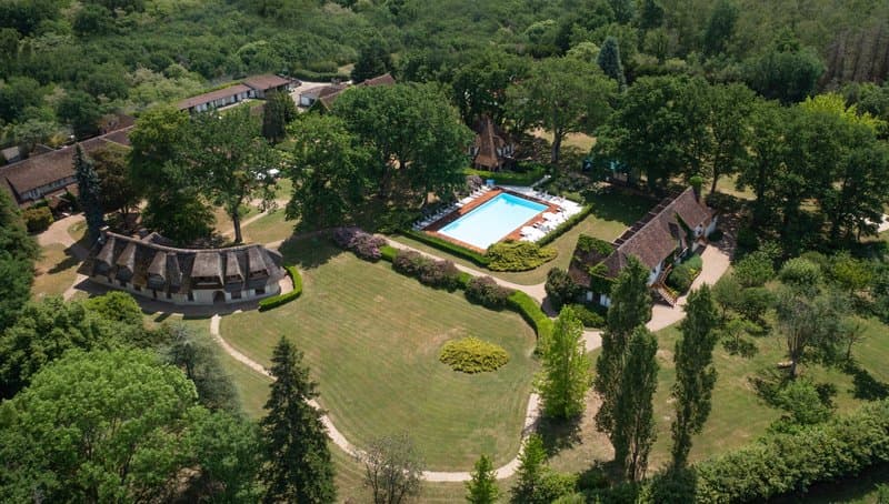 An aerial view of a serene countryside retreat featuring lush greenery, a large swimming pool surrounded by lounge chairs, traditional thatched-roof buildings, and winding pathways, ideal for a tranquil vacation getaway.