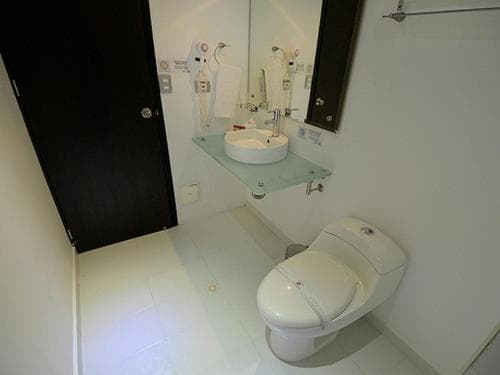 A compact, modern bathroom with white tiled floors, a sleek toilet, a glass shelf holding a sink, and mounted hairdryers offers a clean and functional space ideal for travelers seeking efficient accommodations.