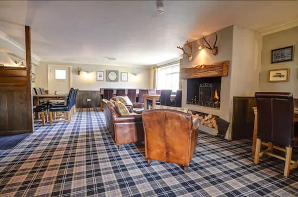 A cozy lodge lounge features leather armchairs, a plaid carpet, a roaring fireplace adorned with antlers, and rustic decor, ideal for travelers seeking warmth and relaxation in a countryside retreat.