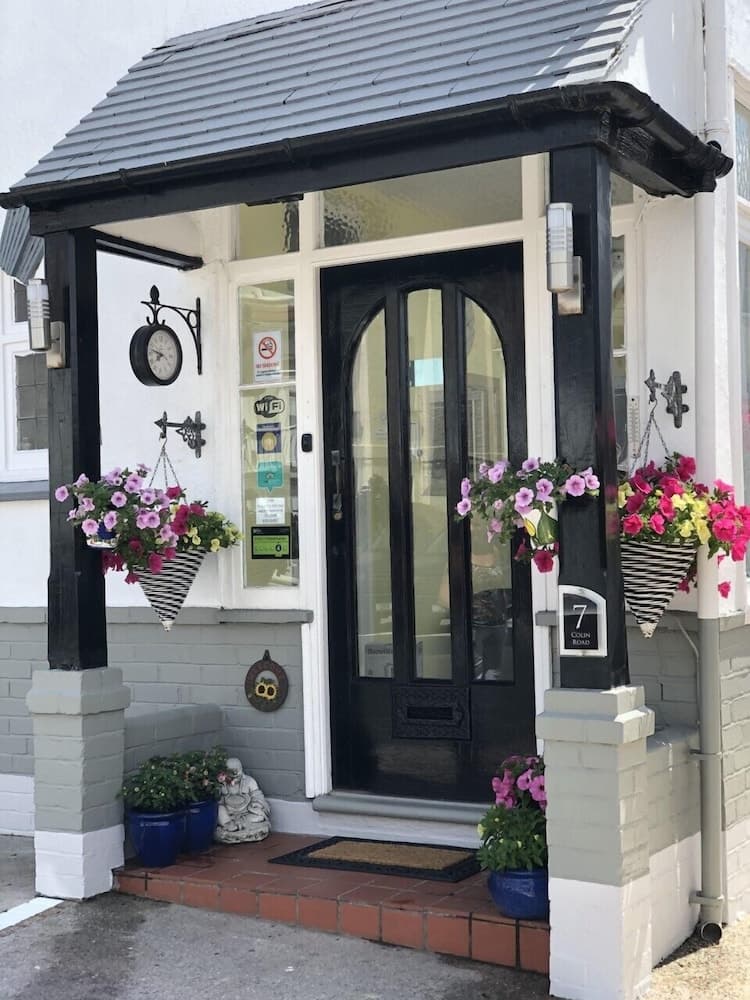 A charming cottage entrance adorned with vibrant flower baskets, a vintage clock, and a cozy doormat invites travelers to explore its quaint and welcoming ambiance.