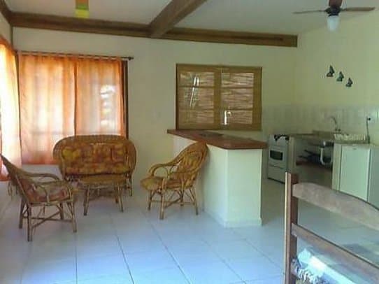 A cozy, sunlit tropical cottage interior features wicker furniture, bamboo blinds, and a compact kitchenette, ideal for travelers seeking a relaxed and affordable accommodation experience.