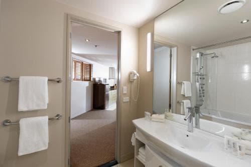 A modern hotel bathroom with a sleek sink, large mirror, and neatly hung towels invites travelers to unwind, with a cozy bedroom visible through an open door in the background.