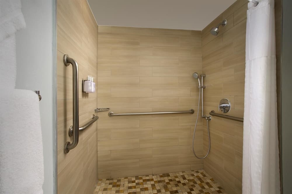A serene hotel bathroom features a spacious walk-in shower with beige tiled walls, safety bars, a handheld showerhead, and a mosaic-tiled floor, ideal for travelers seeking accessibility and comfort.