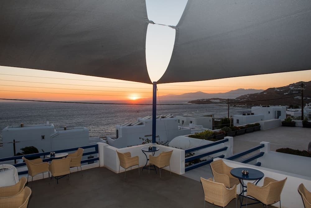 A serene rooftop terrace with cozy seating under a large canopy overlooks the tranquil sea at sunset, ideal for travelers seeking a peaceful retreat in a coastal resort setting.