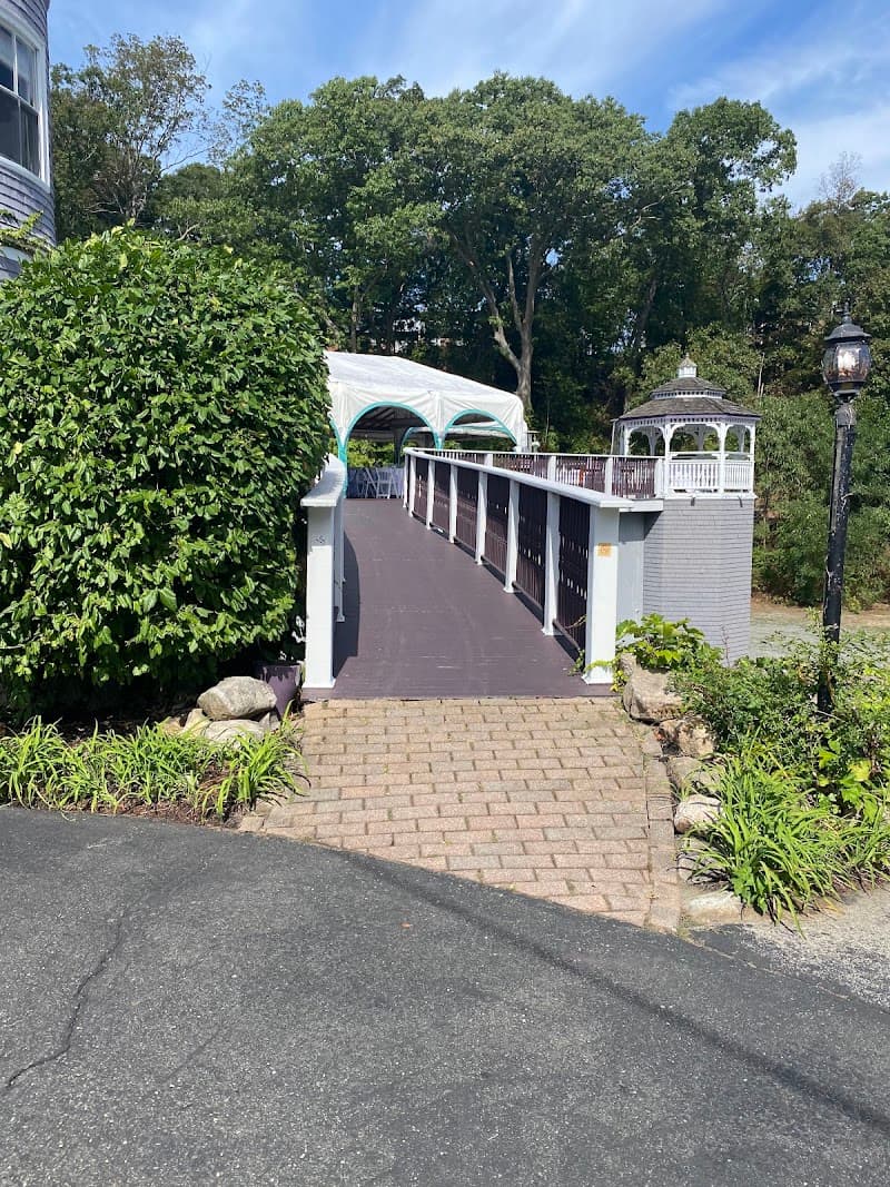 A charming pathway leads to a gazebo and tented area surrounded by lush greenery at the Castle Manor Inn in Gloucester, MA, offering a serene and picturesque setting ideal for travelers seeking relaxation and scenic beauty.