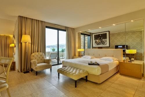 A cozy hotel room with a serene view of the sea through large glass doors, featuring elegant furnishings like a plush bed, armchair, and ottoman, ideal for travelers seeking relaxation and comfort.